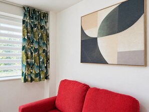Une pièce avec un canapé rouge, une fenêtre avec des stores et un tableau abstrait accroché au mur.