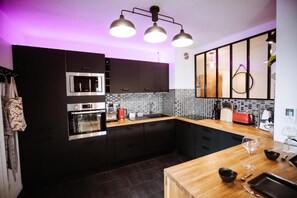 A modern kitchen with dark cabinetry, a built-in oven, a coffee maker, and a wooden countertop. - L'Échappée Parisienne - Canal St-Denis (Aubervilliers)