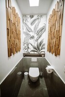 Un baño estrecho con un inodoro, un cubo de basura y un dispensador de papel higiénico. Las paredes presentan un diseño de paneles verticales de madera.