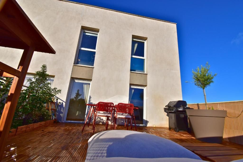 Un edificio moderno con fachada plana, amplios ventanales y una terraza de madera con sillas rojas y una parrilla.