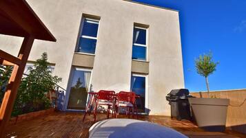 Ein modernes Gebäude mit einer flachen Fassade, großen Fenstern und einer Holzterrasse mit roten Stühlen und einem Grill.