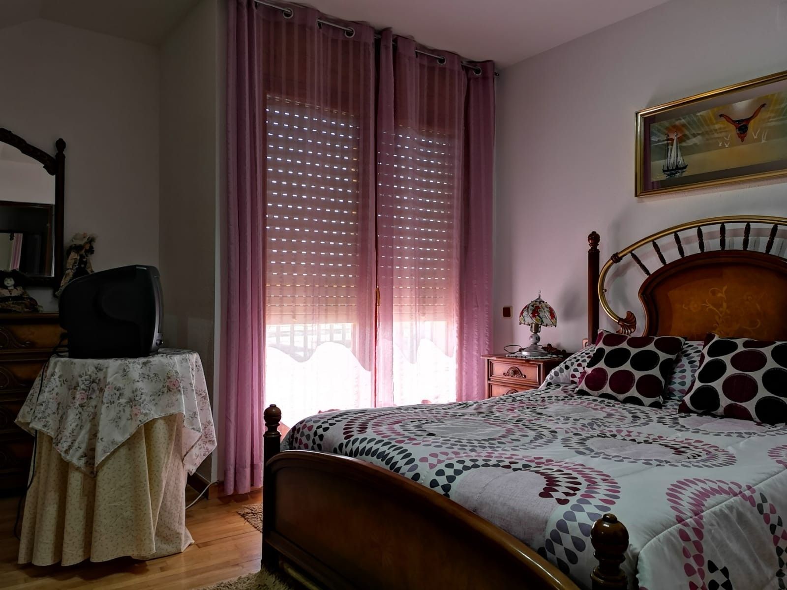 Un dormitorio con una cama de madera, una mesita de noche con una lámpara, una silla y una ventana con cortinas.