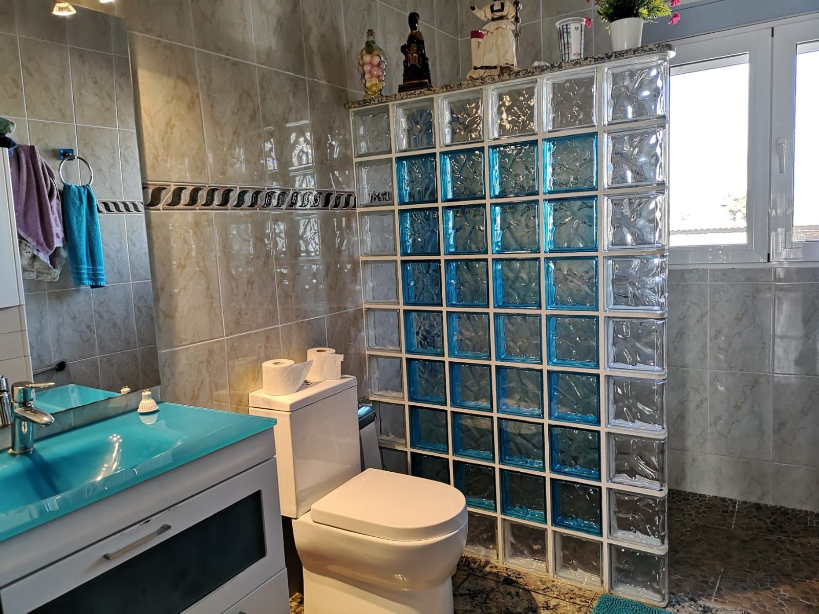Un baño con pared de mármol, cabina de ducha con mampara de vidrio, inodoro blanco y lavamanos azul.