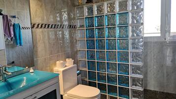 Un baño con pared de mármol, cabina de ducha con mampara de vidrio, inodoro blanco y lavamanos azul.