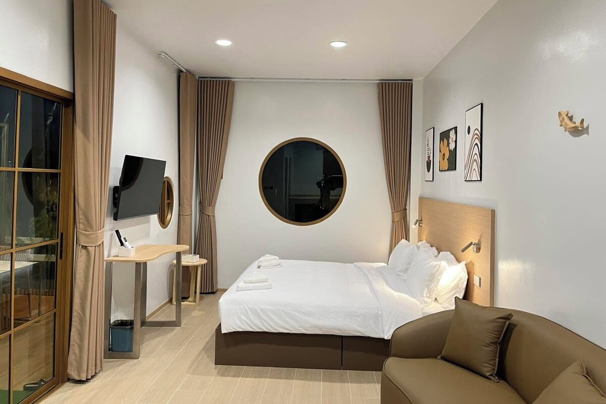 Une chambre d’hôtel moderne équipée d’un lit, d’un miroir rond, d’un bureau avec une chaise, d’une télévision et d’un canapé marron.