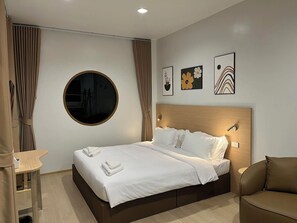 Une chambre d’hôtel avec un miroir rond, un lit recouvert de draps blancs, une tête de lit marron et trois œuvres d’art encadrées accrochées au mur.