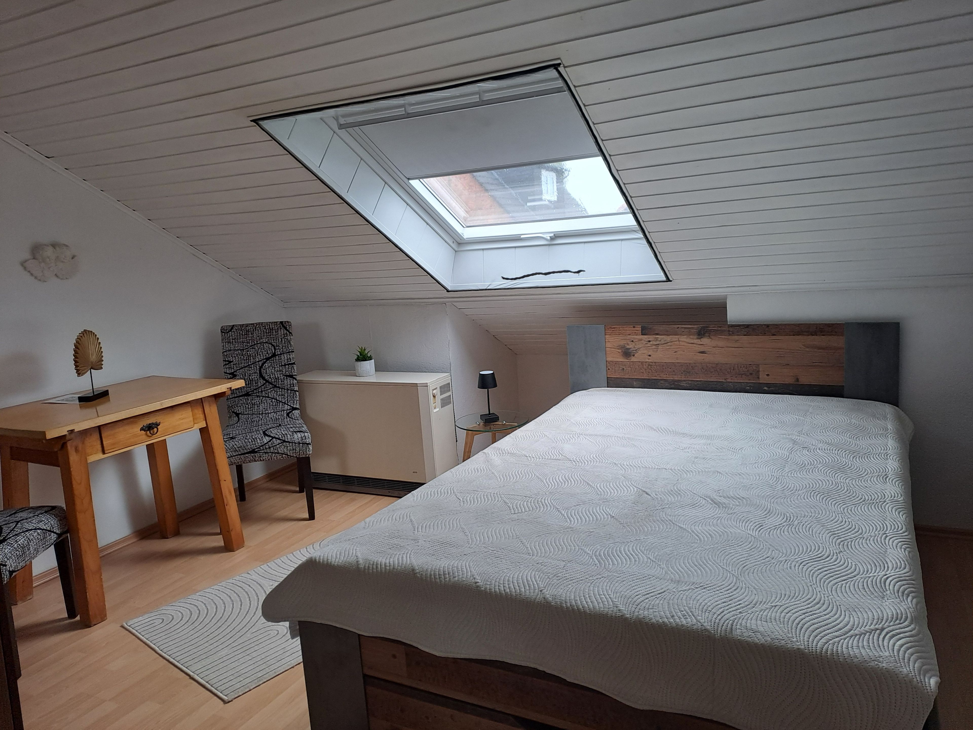 Ein Schlafzimmer mit Dachfenster, einem Bett, einem Schreibtisch, einem Sessel und einem kleinen Tisch mit Lampe.
