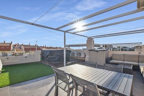 Une terrasse extérieure avec une table et des chaises en bois, un espace salon couvert et une vue sur des immeubles résidentiels en arrière-plan.