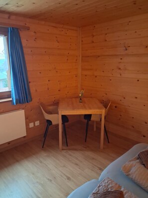 Puinen mökki sisältä, jossa on ruokapöytä ja tuolit, ikkuna verhoineen ja patteri.