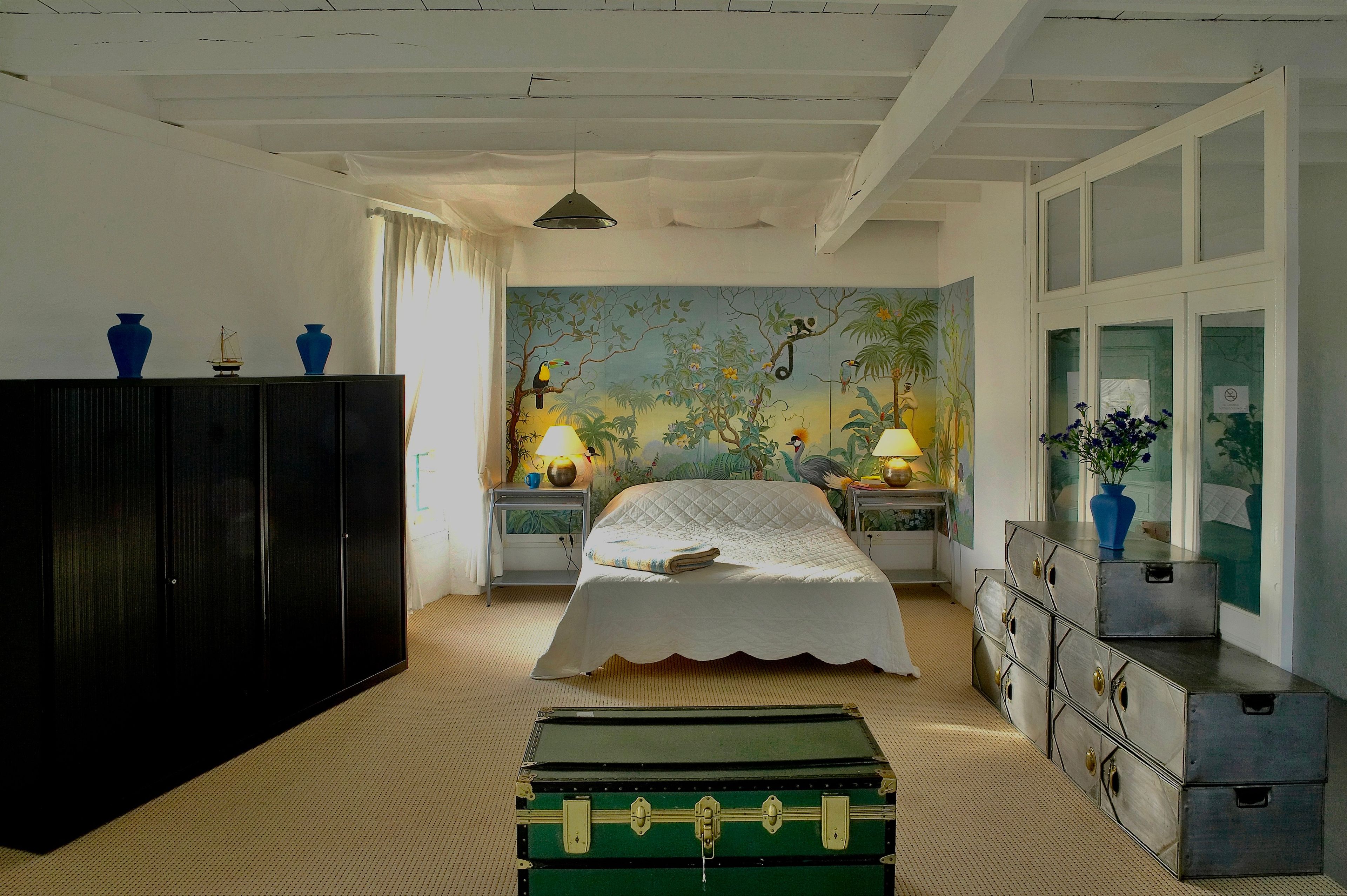 Une chambre à coucher avec un lit, une commode, une table de chevet, une lampe, une valise et une fresque murale.