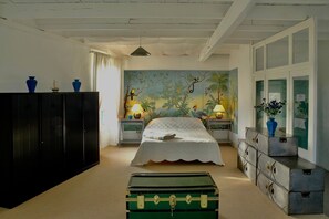 Un dormitorio con cama, cómoda, mesita de noche, lámpara, maleta y un mural en la pared.