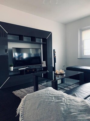 Un soggiorno moderno con una TV a schermo piatto montata su una parete nera, un tavolino da caffè in vetro, un divano nero e una finestra con luce naturale.