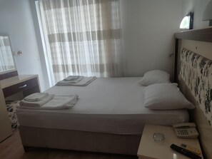 A hotel room with a bed, a bedside table with a phone and remote controls, a mirror, and a window with sheer curtains. - HOTEL YAMUR (ANAMUR)