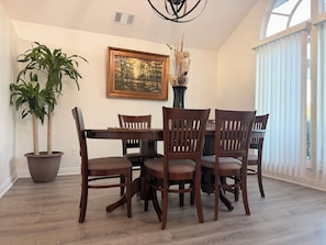 Un comedor con una mesa y sillas de madera, una planta en maceta, un cuadro enmarcado y una ventana con persianas.
