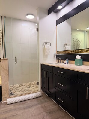 Un baño moderno con ducha envuelta en vidrio, tocador con gabinetes negros y un espejo grande con marco decorativo.