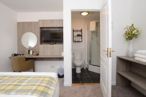 Habitación de hotel con cama, televisor montado en la pared, baño con inodoro y ducha, y una pequeña zona de estar con una silla y mesa.