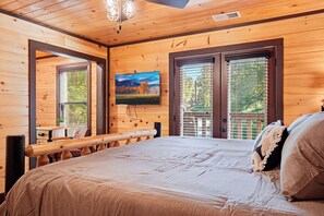 A wooden cabin bedroom with a bed, a television mounted on the wall, and a window with a view of trees. - Spacious Retreat with Hot Tub, Firepit & Easy Access (Sevierville)