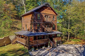 A two-story wooden cabin with a balcony, surrounded by trees. - Spacious Retreat with Hot Tub, Firepit & Easy Access (Sevierville)