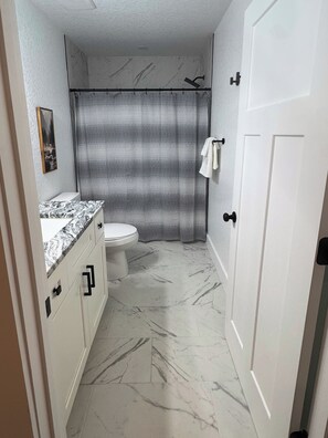 A bathroom with a toilet, a shower with a patterned curtain, marble flooring, and a door with a handle. - Highlands Time - New 3BR Home 1 Mile to Glen Falls, 5 Min to Downtown Highlands (Highlands)