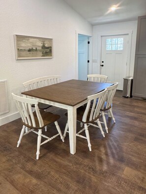 A dining area with a wooden table and white chairs, a painting on the wall, and a door with a window. - Highlands Time - New 3BR Home 1 Mile to Glen Falls, 5 Min to Downtown Highlands (Highlands)