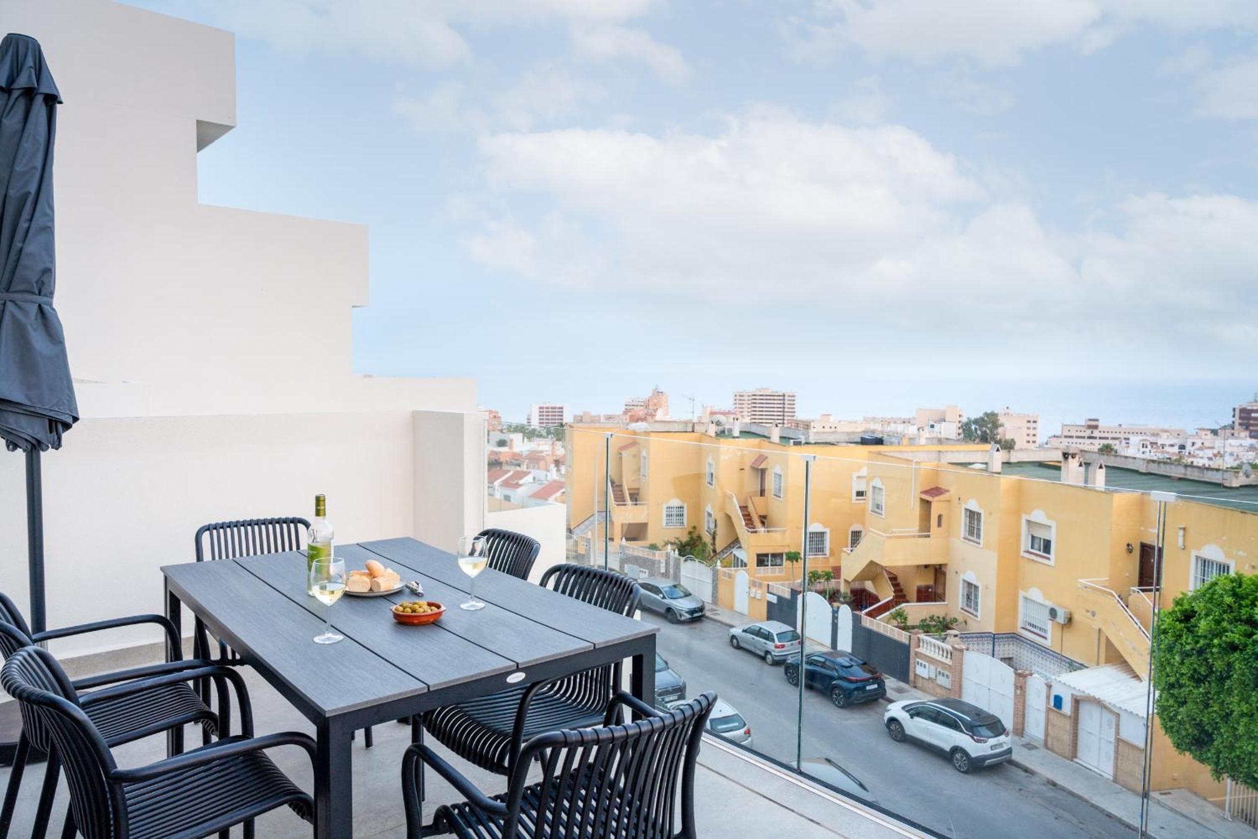A balcony with a table and chairs, overlooking a street with parked cars and buildings.
