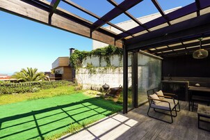 Un espacio exterior moderno con una pérgola de madera, un ventanal que da a un área interior con mesa y sillas, y un césped.