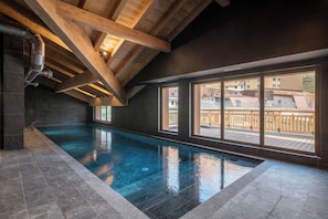 Une piscine intérieure donnant sur une terrasse en bois et des bâtiments situés à l’extérieur.