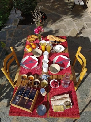 Un coin pique-nique aménagé en plein air, avec une table dressée pour deux personnes, comprenant des assiettes, des couverts, un panier de pain, des fruits et une bouteille de jus.
