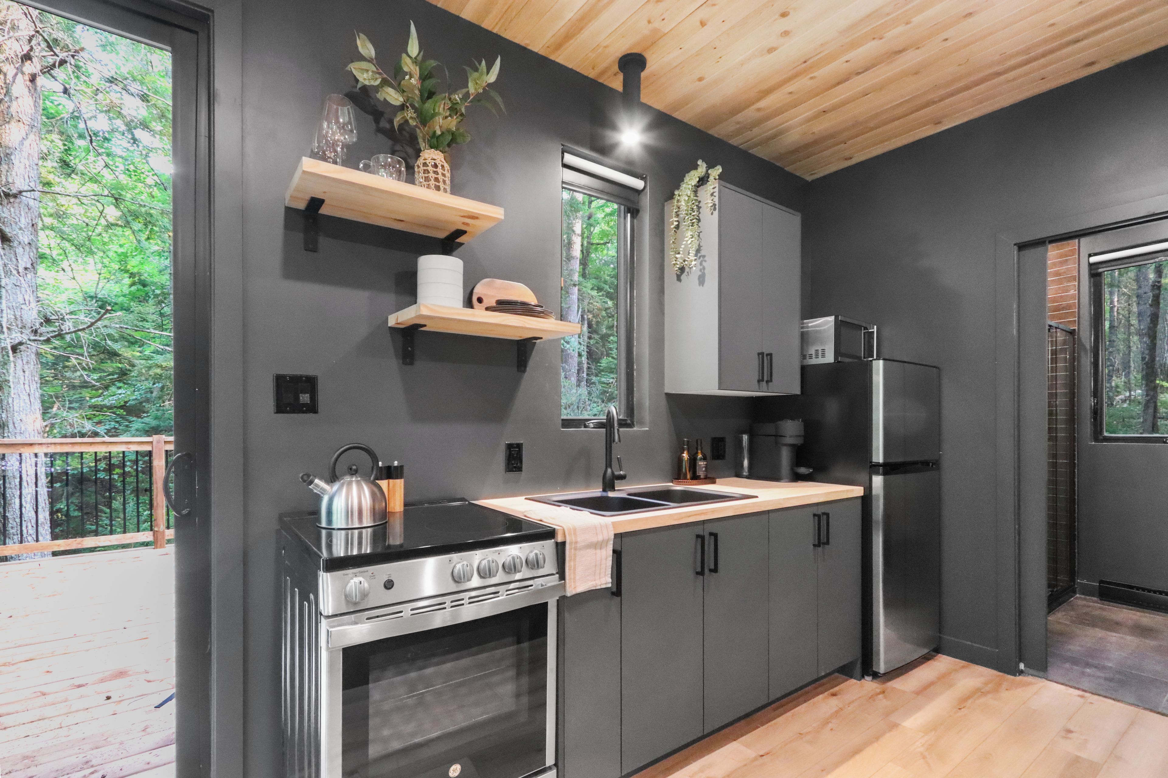 Une cuisine moderne équipée d’un four, d’une cuisinière et d’un réfrigérateur encastrés. On y trouve des étagères en bois au-dessus de l’évier et une fenêtre donnant sur un paysage verdoyant.