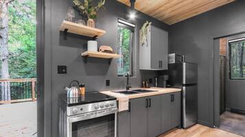 Une cuisine moderne équipée d’un four, d’une cuisinière et d’un réfrigérateur encastrés. On y trouve des étagères en bois au-dessus de l’évier et une fenêtre donnant sur un paysage verdoyant.