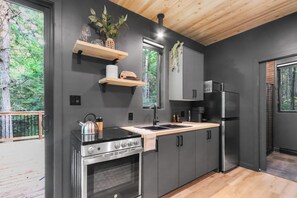 A modern kitchen with a built-in oven, stove, and refrigerator. There are wooden shelves above the sink and a window with a view of greenery. - La P'tite Maison - On the Richibucto River (Weldford)