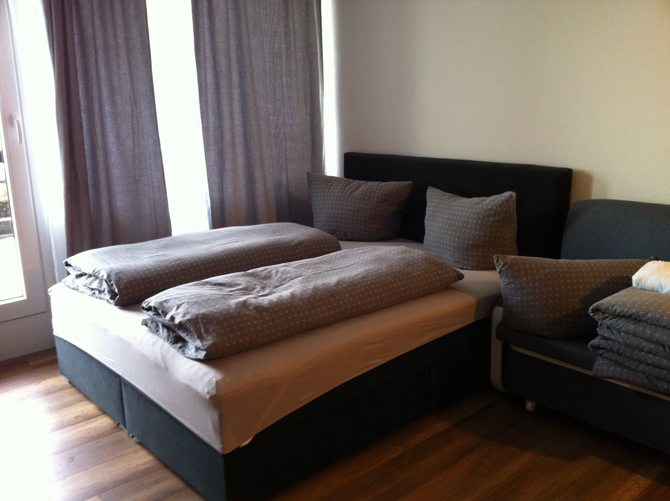 Ein Doppelbett mit dunklem Kopfteil, zwei Kissen und einer Decke. Rechts daneben befindet sich eine Couch mit Kissen und Decke.