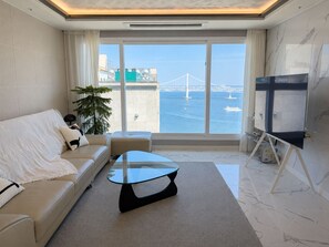 Un salon moderne avec un canapé, une table basse en verre, un téléviseur et une grande fenêtre donnant sur un pont et l’eau.