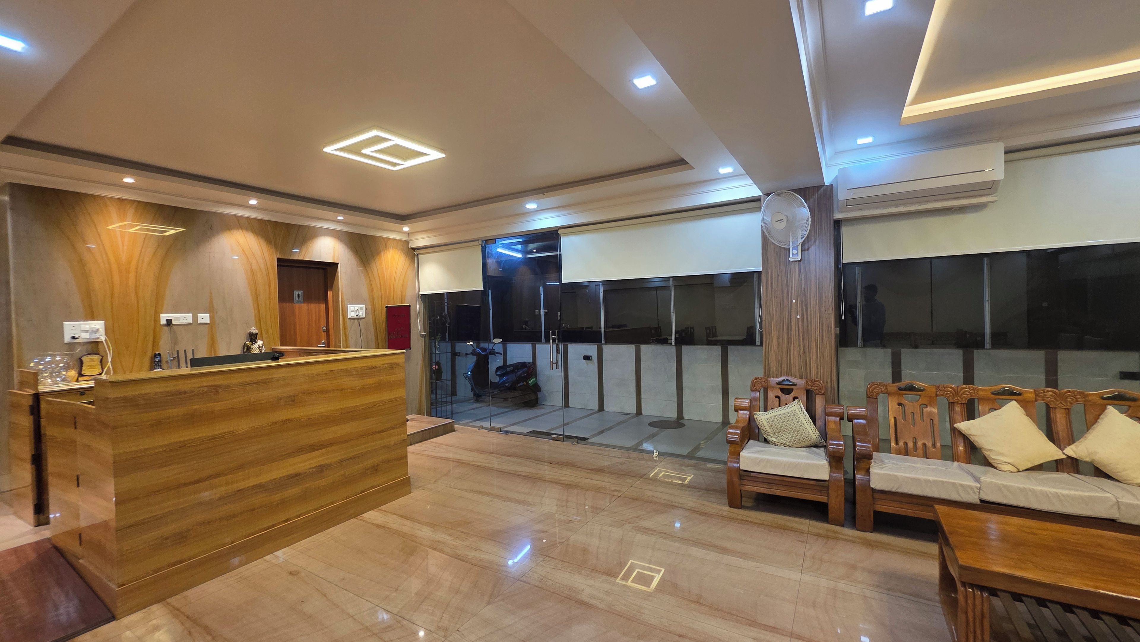 A hotel reception area with a wooden reception desk, a seating area with wooden chairs, and a glass partition.