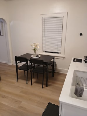 Uma área de refeições com mesa e cadeiras, uma janela com persianas e uma bancada branca com pia.