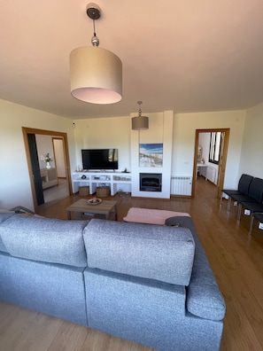 Una sala de estar moderna con un sofá gris grande, un televisor de pantalla plana, una chimenea y una zona de comedor con una mesa y sillas de madera.