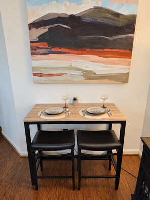 A dining area with a table set for two, two black chairs, and a large abstract painting on the wall. - Heart of Wintergreen 1BR - Fully Stocked, Year-Round, Couples & Families (Wintergreen Resort)