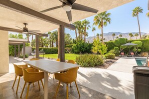 An outdoor patio with a table and chairs, a ceiling fan, and a view of a pool and mountains. - Palm Springs Oasis | Private Pool, Spa & Game Room (Palm Springs)