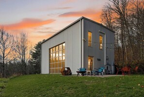 A modern two-story building with a large garage door, outdoor seating area with chairs, and a view of the sunset. - The Looking Glass Modern Vermont Retreat with Private Hot Tub (Barnard)