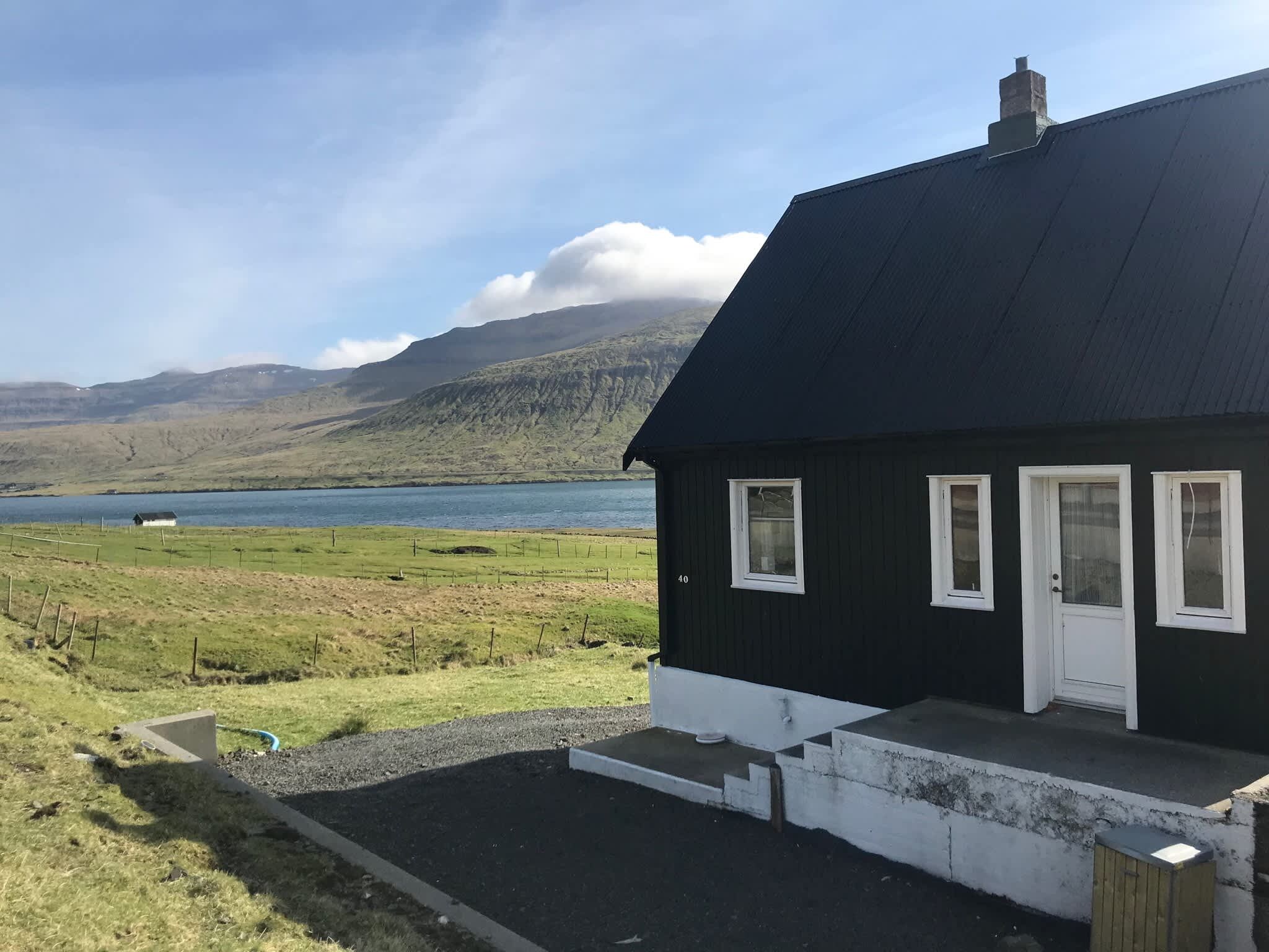 A black house with a white door and windows, a gravel driveway, and a view of a lake and mountains in the background.