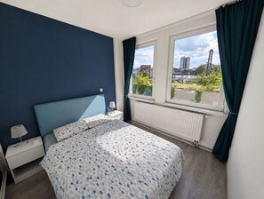 Ein Schlafzimmer mit einem Bett, einem Nachttisch, einem Fenster mit Vorhängen und Blick auf die Stadt.