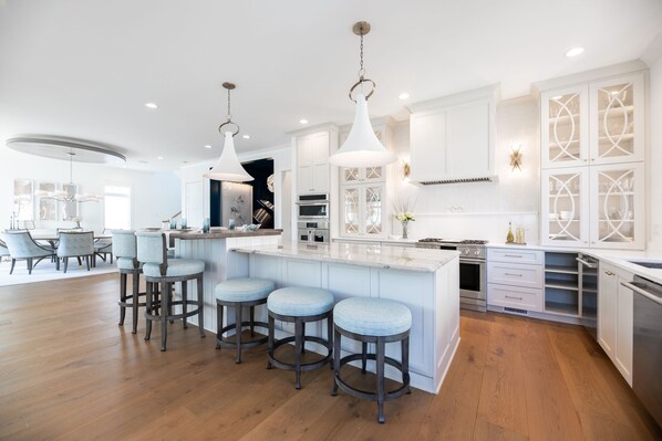 A modern kitchen with white cabinetry, a central island with bar stools, and stainless steel appliances. - Luxury Coastal Estate + Ocean & ICW View + Private Pool + Grand Piano & Wine Bar (Ocean Isle Beach)