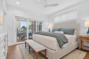 A bedroom with a large bed, a bench, a ceiling fan, and a view of the outdoors. - Luxury Coastal Estate + Ocean & ICW View + Private Pool + Grand Piano & Wine Bar (Ocean Isle Beach)