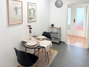 Un coin repas avec une table dressée pour deux personnes, une horloge murale et une étagère ornée d’une plante.