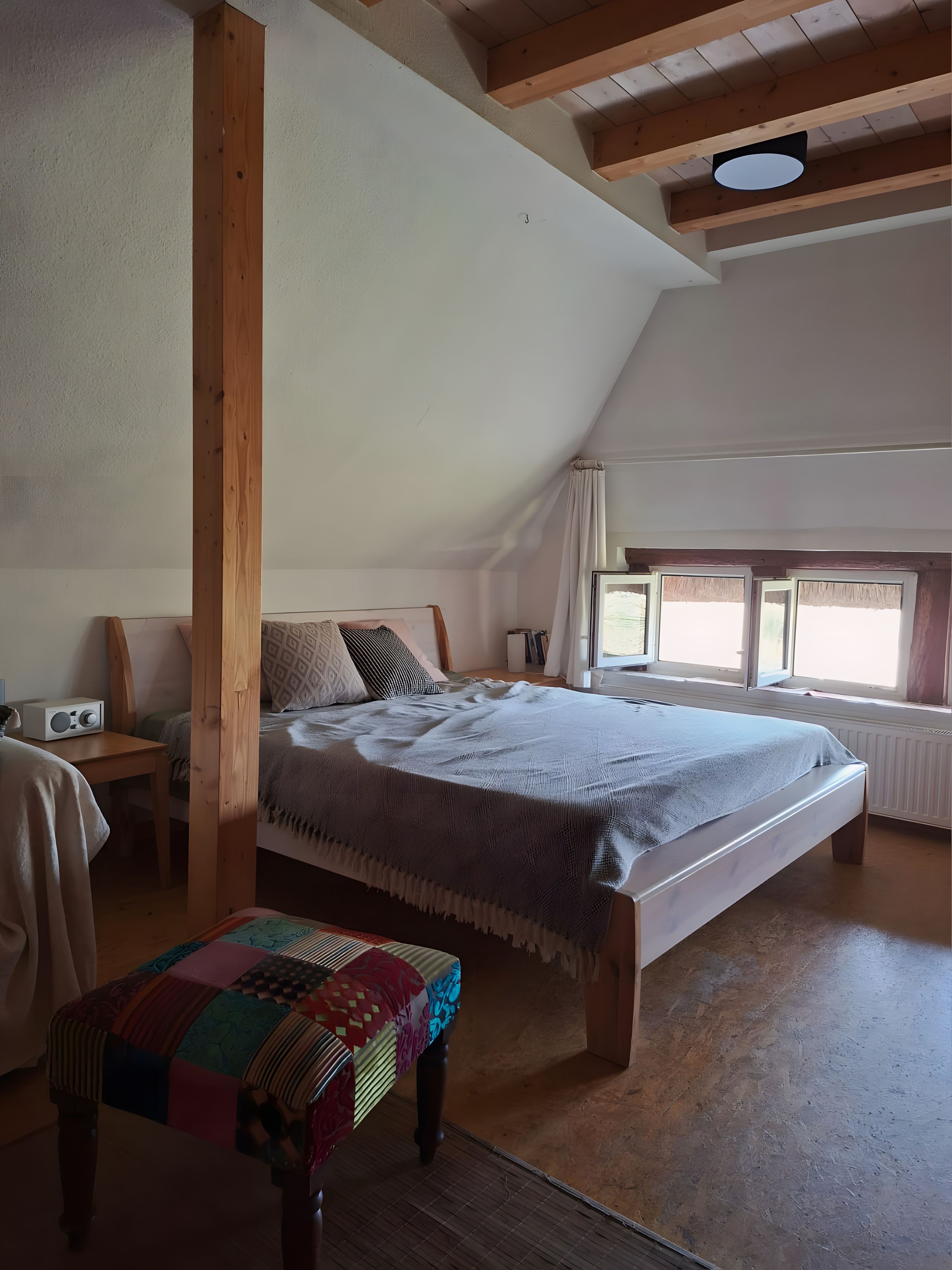 Een slaapkamer met een schuine balkenkap, een bed met een beddegoed met patroon, een houten bankje en een raam met gordijnen.