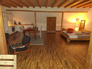 Um quarto acolhedor com piso de madeira, uma cama com almofadas, uma mesa com cadeiras e uma cozinha.