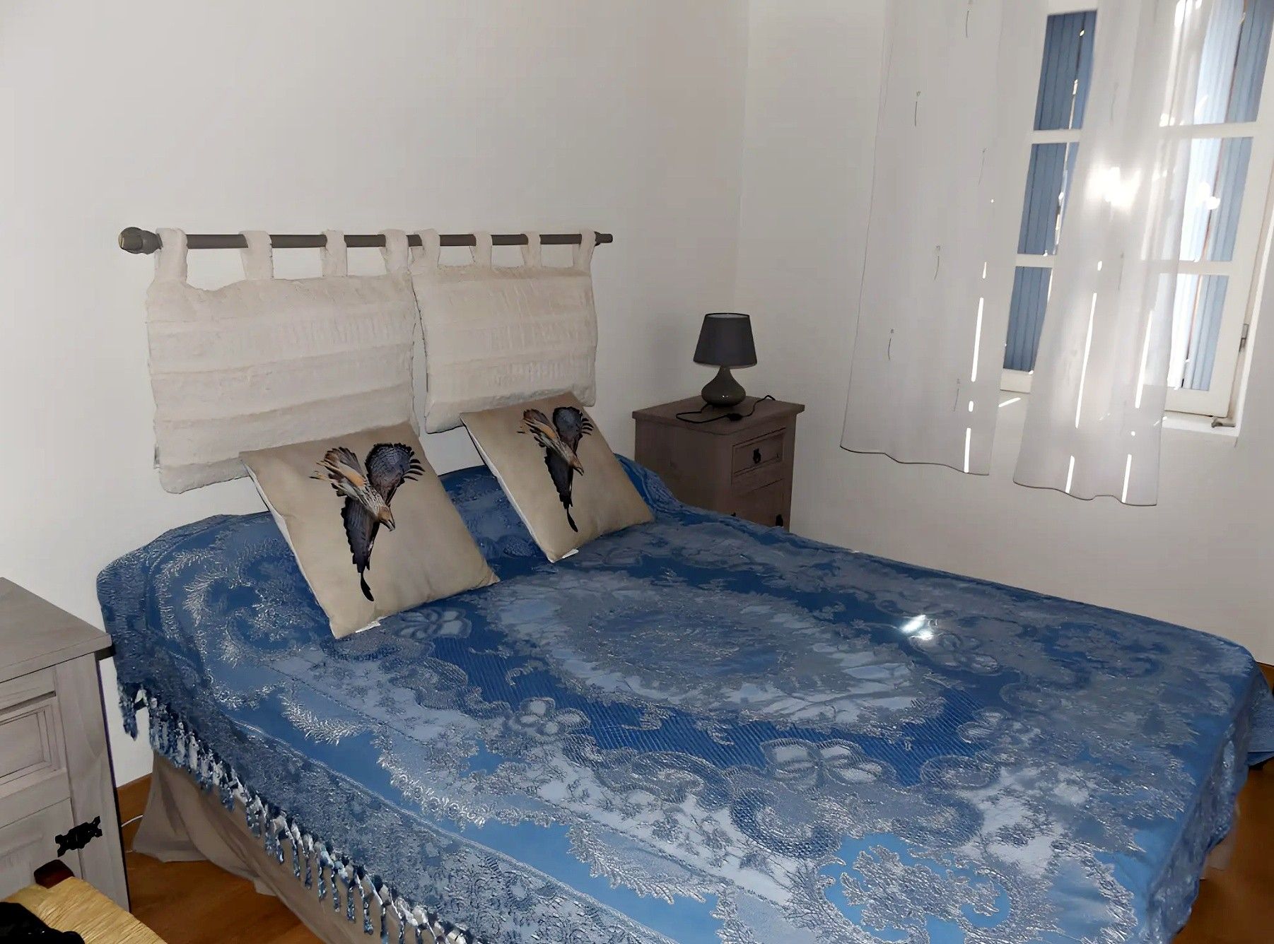 A bedroom with a bed, pillows, a nightstand, a lamp, and a window with curtains.