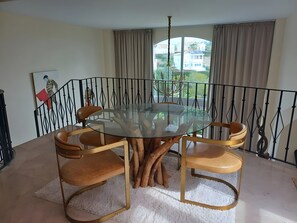 Un coin repas avec une table ronde en verre entourée de chaises, un tapis blanc à texture particulière et une vue sur les bâtiments à travers une grande fenêtre ornée de rideaux.