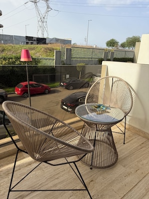 A balcony with wicker chairs and a glass table, a red car, and a black car parked in the background. - New Listing (Casablanca)