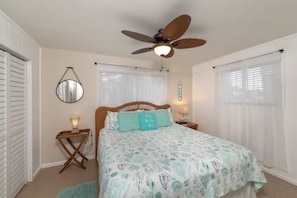 Quarto com cama, ventilador de teto, mesa de cabeceira com abajur e janela com persianas.